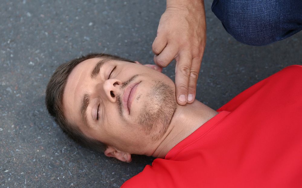 White man in red shirt knocked out on sidewalk with someone putting two fingers on neck to feel for pulse