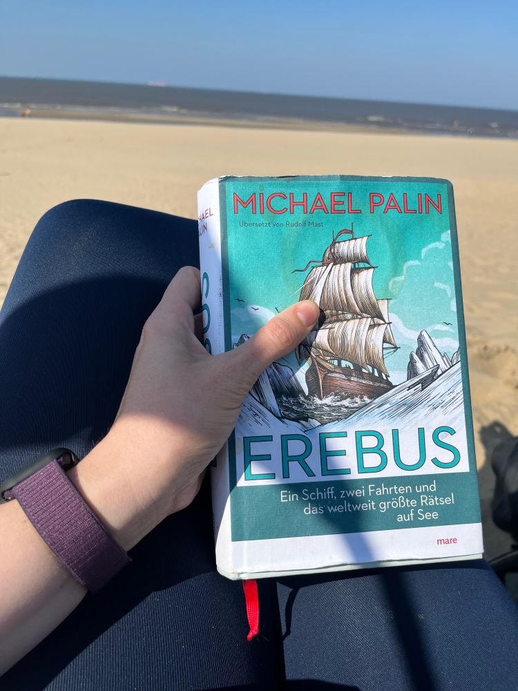 Das Buch “Erebus” von Michael Palin in der deutschen Ausgabe wird in die Kamera gehalte. Im Hintergrund eine Strandkulisse.