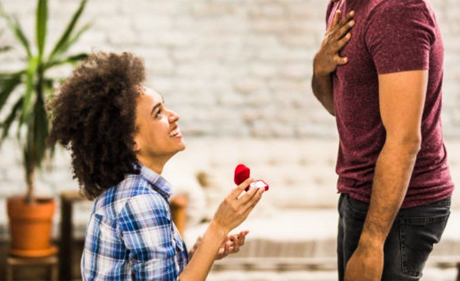 light-skinned black woman with an afro, in a plaid shirt. she is on bended knee with a red ring box in her hand, proposing to man standing before her. he is wearing a fuchsia shirt and black jeans