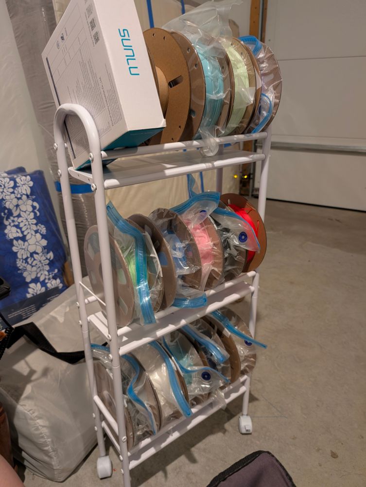 A white pipe frame shelf in a garage. It's holding spools of 3d printer filament in resealable bags.