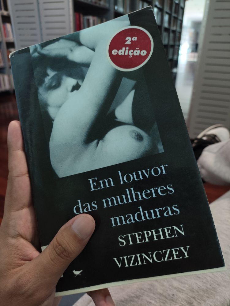 Foto do livro "Em louvor das mulheres maduras" de Stephen Vizinczey. Ao fundo aparecem estantes de livros da biblioteca. 