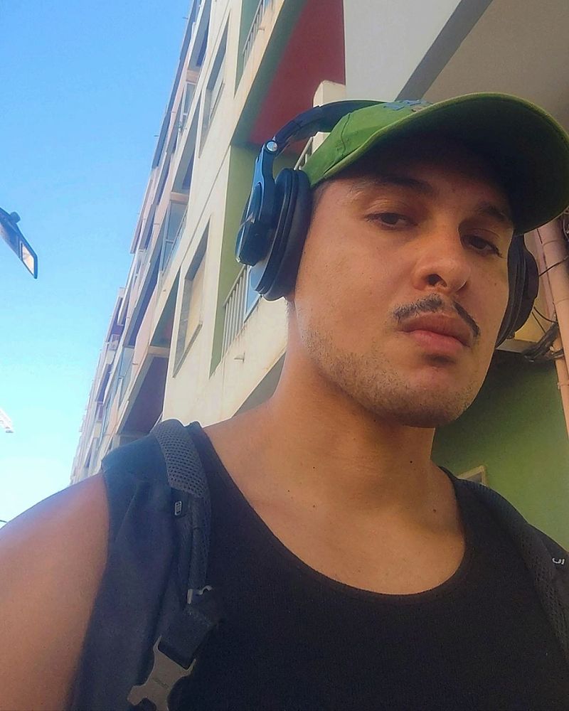 Self de baixo para cima, estou de boné verde, headphone, regata preta e as alças de uma mochila preta aparecem. Sou um homem pardo de bigode.