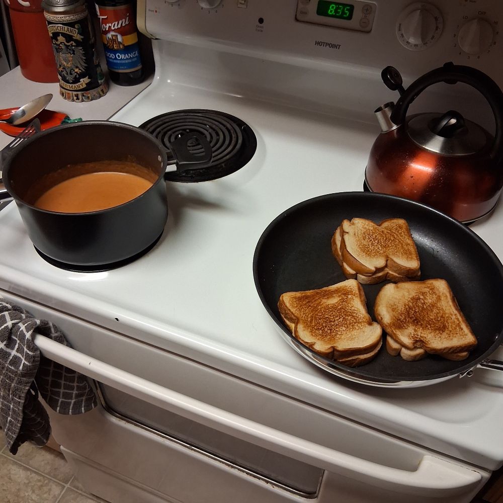 Grilled cheese sandwiches cooking in a pan, a pot with tomato soup next to it
