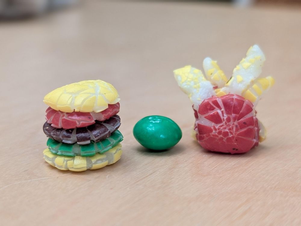 A macro portrait photo of small multi-colored Skittles candies on a laminated tabletop which have been squished, reshaped, and combined into roughly the shape of a fast-food hamburger and a side of french fries.