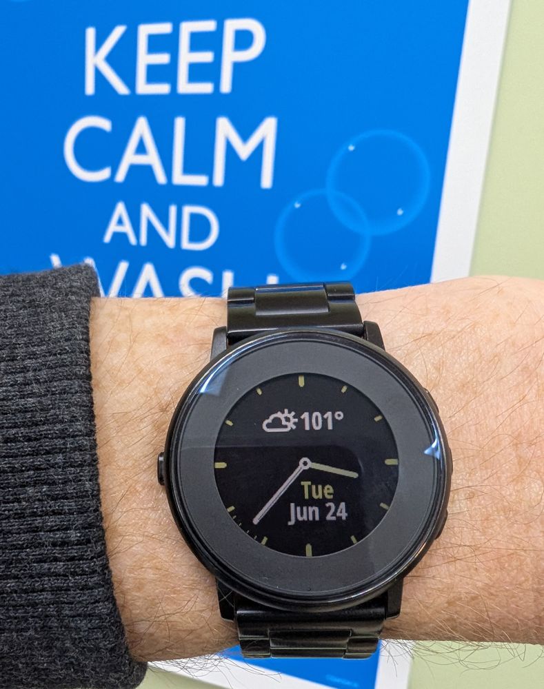 Photo of a Pebble Time Round smartwatch being worn, sporting the "Obsidian" analogue-style watchface, which indicates the time (15:37), day, date, temperature (101°F) and conditions (partly cloudy). Part of a wall poster can be seen in the background, which reads "KEEP CALM AND WASH YOUR HANDS".