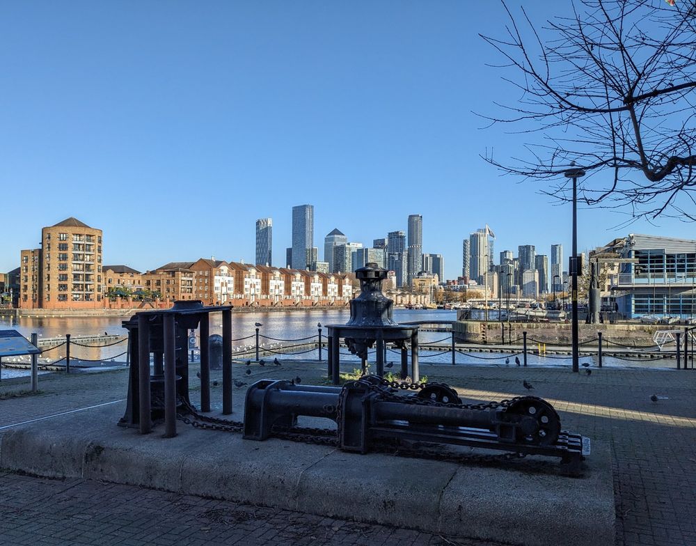 Disused dock (Greenland Dock) surrounded by housing with old machinery in the foreground and tower blocks of Canary Wharf in the background against a clear blue sky.
