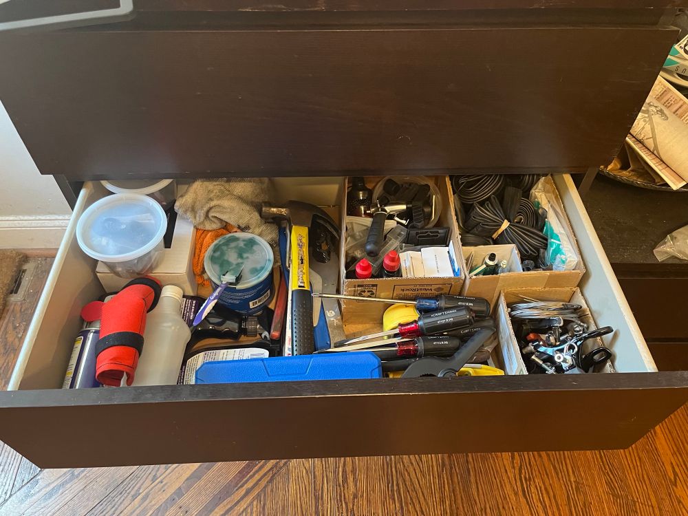 A drawer in an aging IKEA dresser, set next to my front door, with a bunch of bicycle-relevant parts and tools. Enumerating every item here would be exhausting, but, organizationally: • a couple of half-quart containers sit at the top left, atop a carbon fiber repair kit • in front of those are “fluids” (Simple Green, WD-40, isopropyl alcohol) • to the right are rags and a pot of grease (for axles, cranks, seat posts, etc) • to the right of that is a box holding mostly “chain stuff” (links, extra chains, chain lube, a chain break tool that is human-hand-sized, rather than the one in the multitool you’ve used to do that before) • to the right of that is all tube/tire stuff (tubes, segments of tubes, several Presta and Schrader valves clipped out of burst tubes awaiting part component extraction if necessary) • looping clockwise, the next bin is brake stuff (cables, pads, a couple sets of calipers) • left of that is the pile of “short tools”: clamps, small wrenches, screw drivers, measuring tape, fluid level • left of that is the (in a case) digital caliper atop the “long tools” (wrenches, chain whip, hammer)