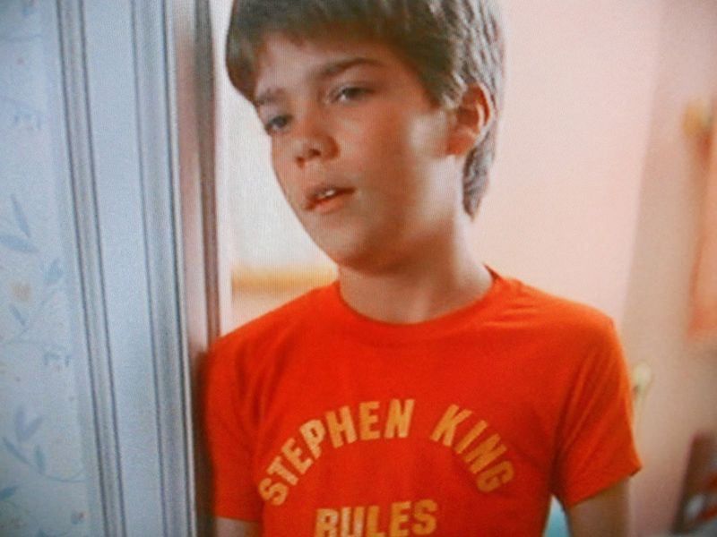 A stereotypical late 1970s boy wearing a red t-shirt emblazoned, as if for a sports team, in yellow with, “Stephen King rules”.