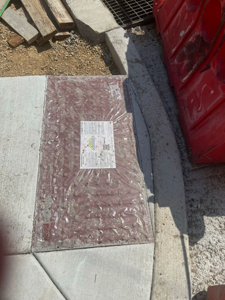 A red-grey textured panel set into newly-poured cement at a curb cut for pedestrian crossing in South Philly.

It is so recently installed that the plastic covering it ships in is still attached.