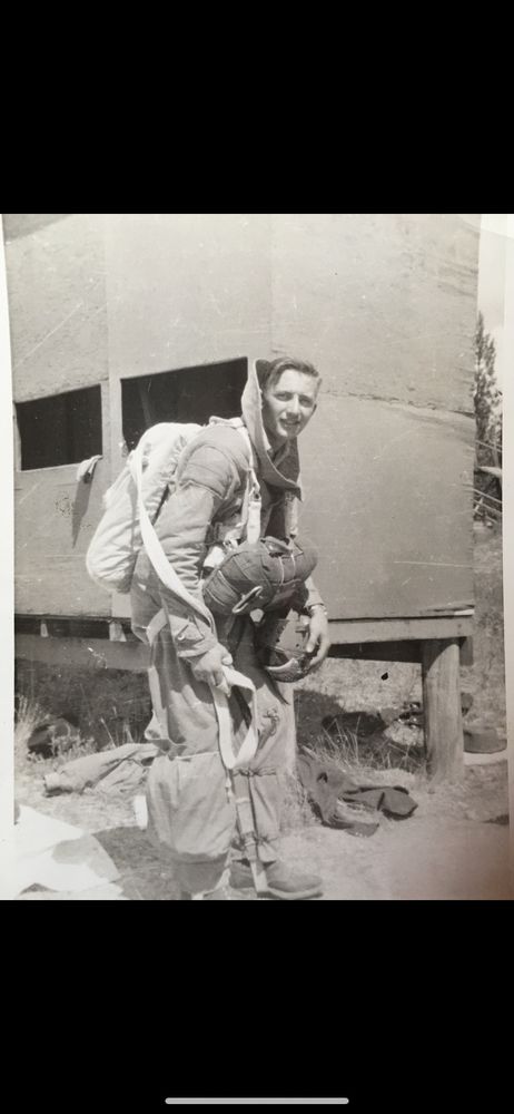 A black and white photograph of a young white man in his Smokejumping gear, including the parachute he would use to parachute into inaccessible wilderness to fight fires.