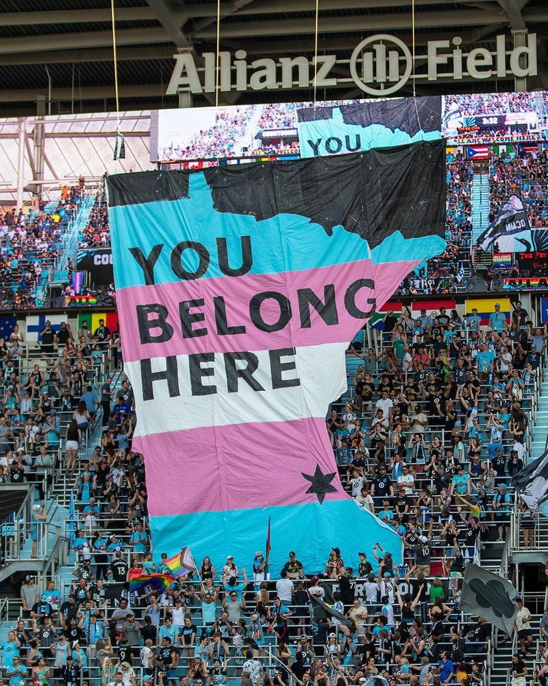 A large banner depicting the state of Minnesota in the colors of the transgender flag and the words "You Belong Here" is displayed above the supporters section at Allianz Field.