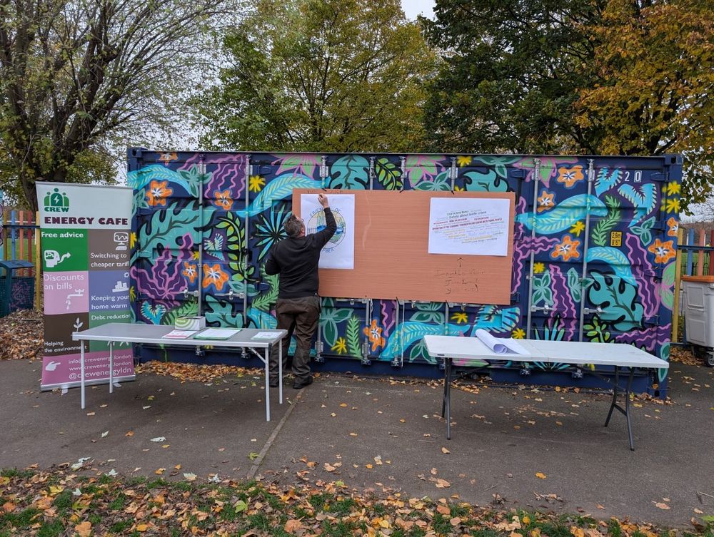 A man is in a park with his back to the camera. He pins some posters to a container covered in street-art style art in blue, purples and greens. To the left is a CREW Energy Cafe pull-up banner. In front of the container there are two tables with papers and leaflets on top.