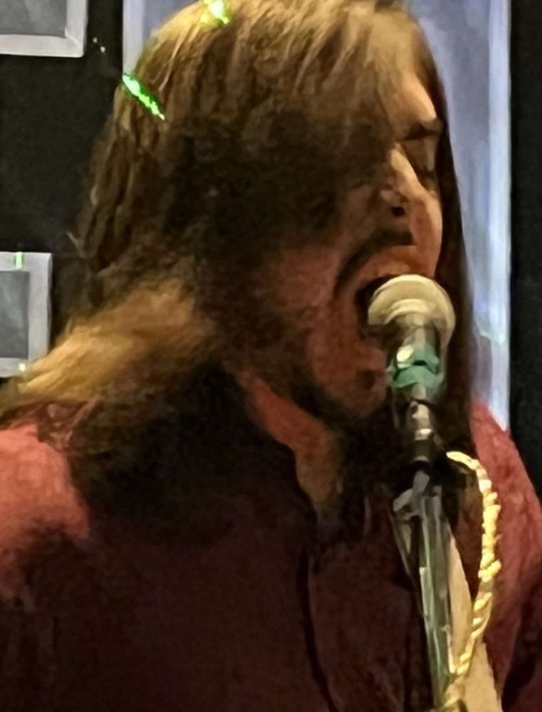 me in a burgundy button up shirt violently yelling into a microphone with a white guitar strap on my left shoulder (the guitar is cropped out of the photo)