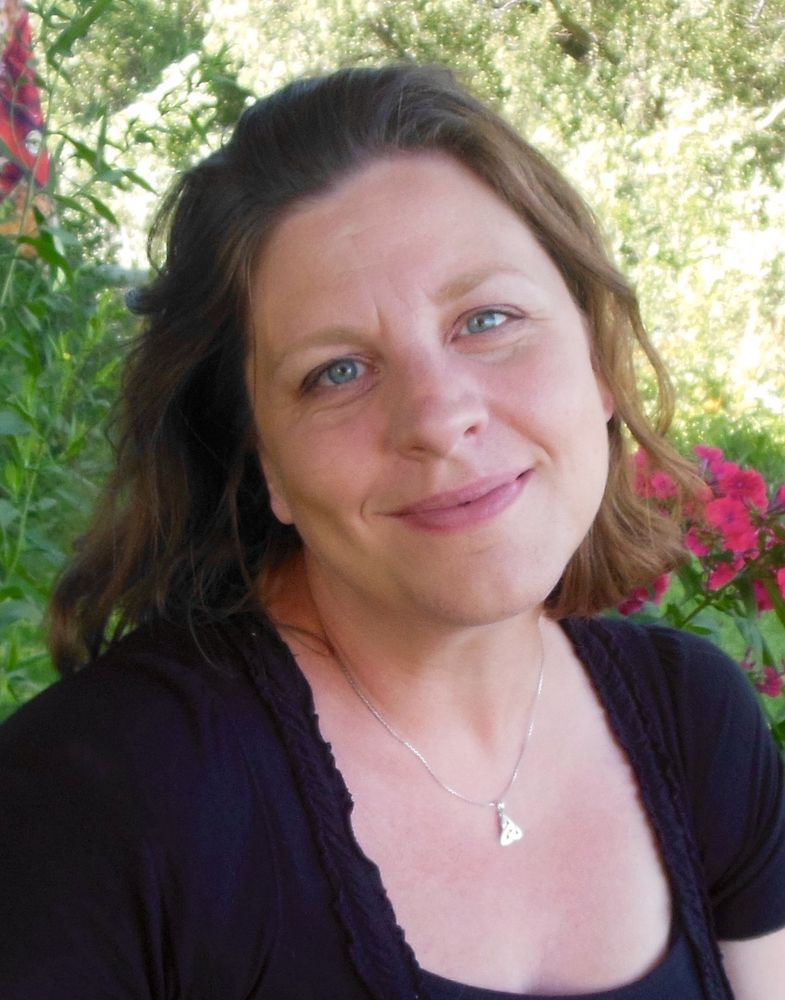 Picture of me, Carye VanDerPol Mahoney: white woman with shoulder length wavy brown hair, blue/green eyes and a warm smile, head slightly cocked to the side, wearing a black shirt and a silver necklace with a triquetra symbol. There is greenery and magenta flowers in the background