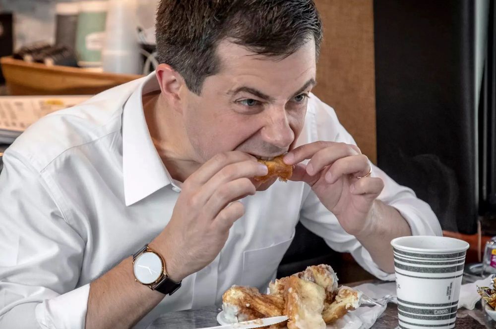 Pete Buttigieg is pictured holding a piece of a cinnamon roll in both hands as he bites the center of it. Below is a plate with the other remnants of a cinnamon roll, but cut apart in a way that would be considered somewhat unusual for someone to eat a cinnamon roll.