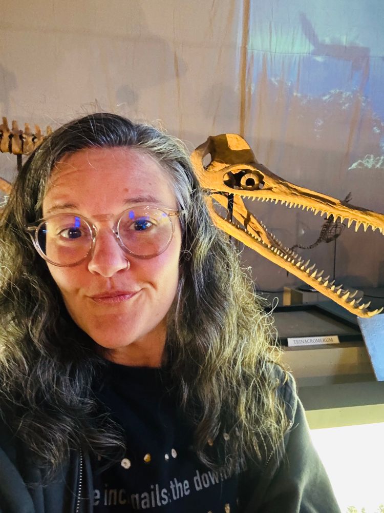 A selfie of a person with long wavy hair and glasses and a Plesiosaur skull behind them. The skull is posed with jaws open so there are many Plesiosaur teeth visible 