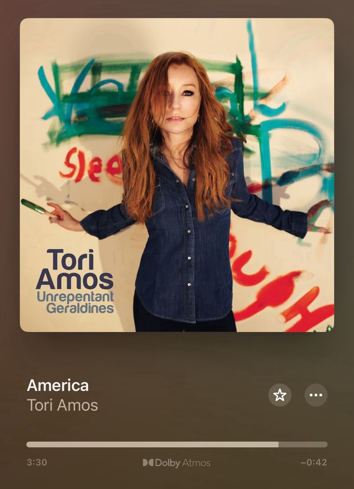 A screenshot of apple music- tori amos' album Unrepentant geraldines is playing mostly through the first song, America. The album cover shows Amos, a woman with long red hair in a blue denim shirt, standing in front of painted words.