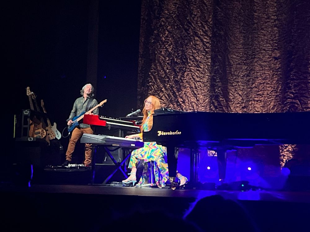 An image from a San Francisco 2023 Tori Amos concert. Tori is at the keyboard with a bosendorfer in front of her and is wearing a rainbow-striped outfit. To her right is Jon Evans playing bass.
