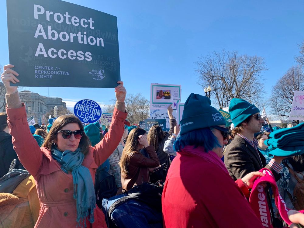 A woman in a pink coat and teal scarf stands in a crowd at a rally, and hold a teal sign that reads “protect abortion access.”