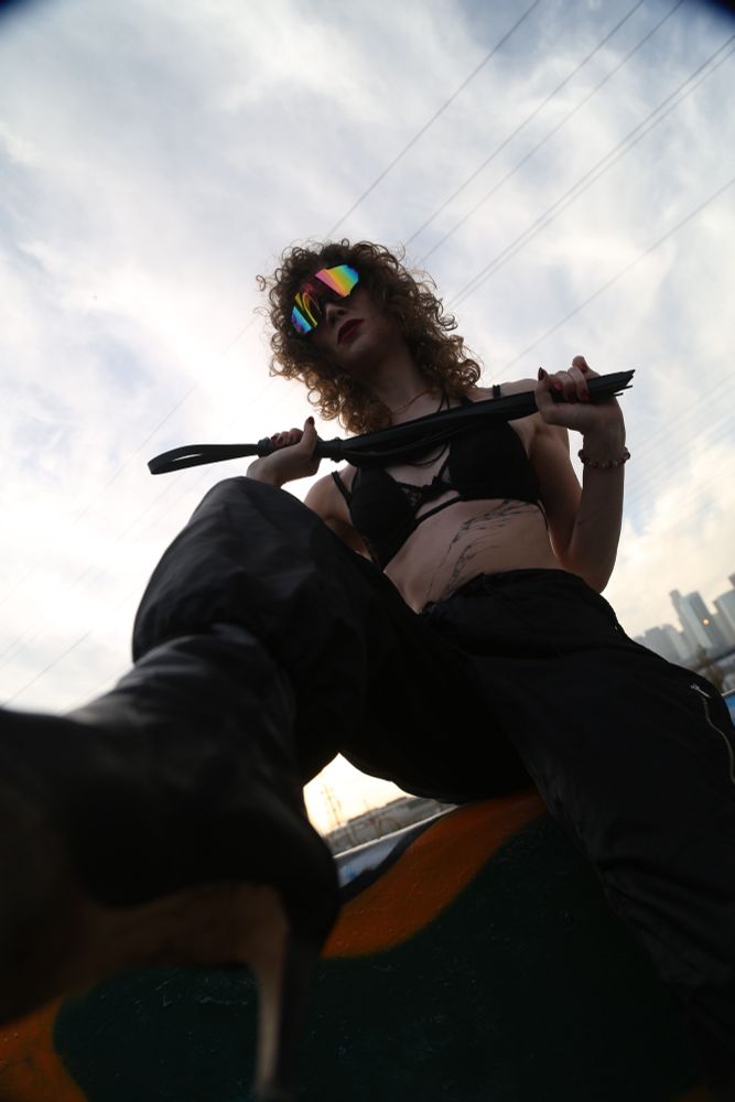 Goddess Ivy Stone, viewed from below, in stiletto boots, black combat pants, and a black bra, holding a flogger
