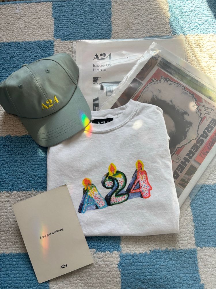 A24 birthday lit candle logo T-shirt (white, logo is colorful), light blue logo hat, and two zines (the covers show Eraserhead on one, the other has a small White House), and a note that says, “enjoy your special day” Background, a blue and white checkered area rug. 