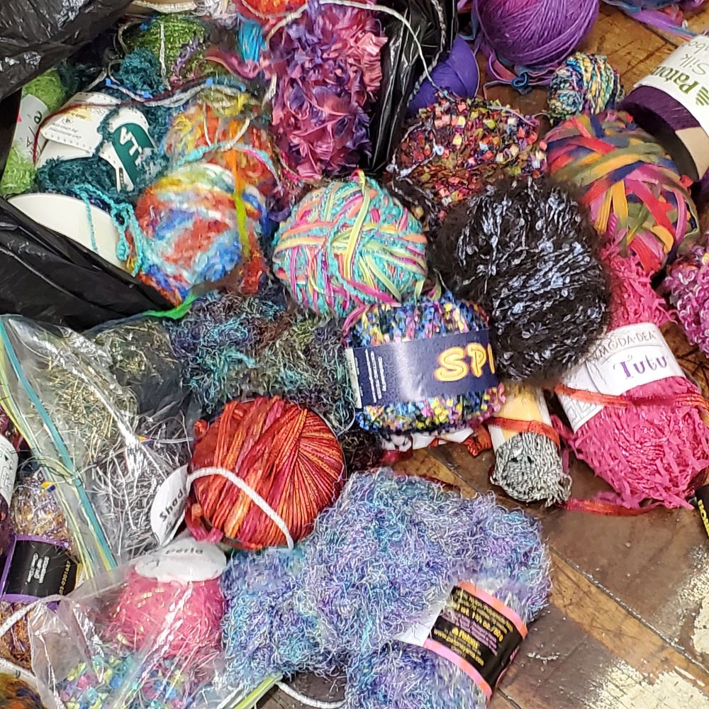 A pile of yarn balls in bright colors tumbling out of a bag, purple, pink, orange, rainbow mixes, fuzzy, furry, metallic blends.