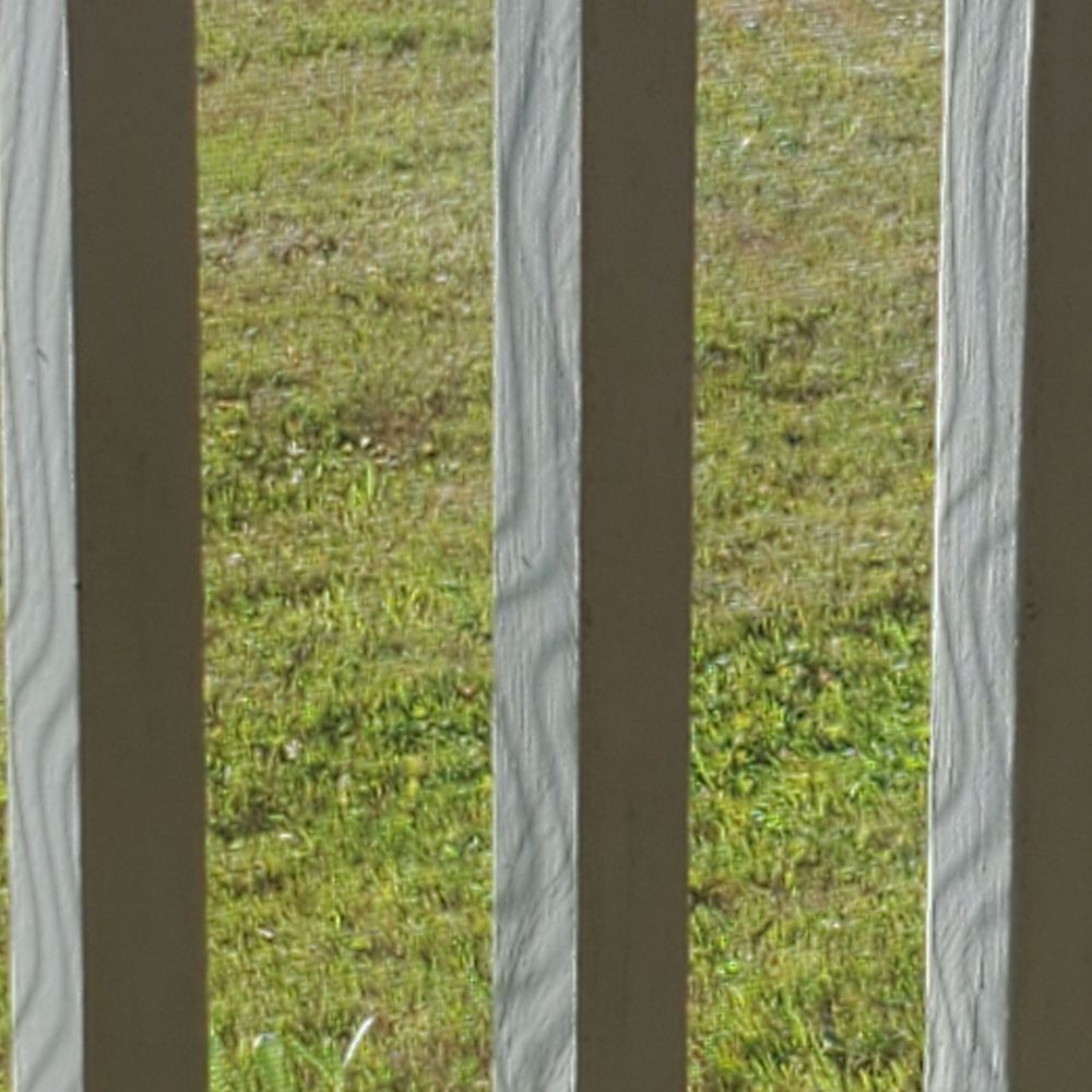 Moire shadows or sunlight through a screen on railing posts, with grass in the background. 