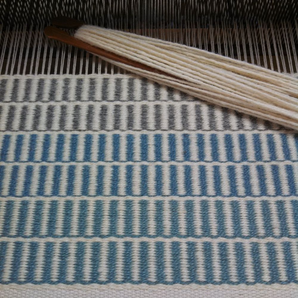 Wool Rug I made, in progress, on a Hand Loom with Stick Shuttle. Horizontal rows of blue and gray vertical stripes alternating with natural white. 