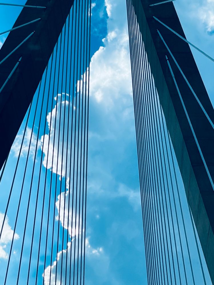 photo of suspension wires going up into the frame of a bridge into charleston, with white clouds and blue sky in the background