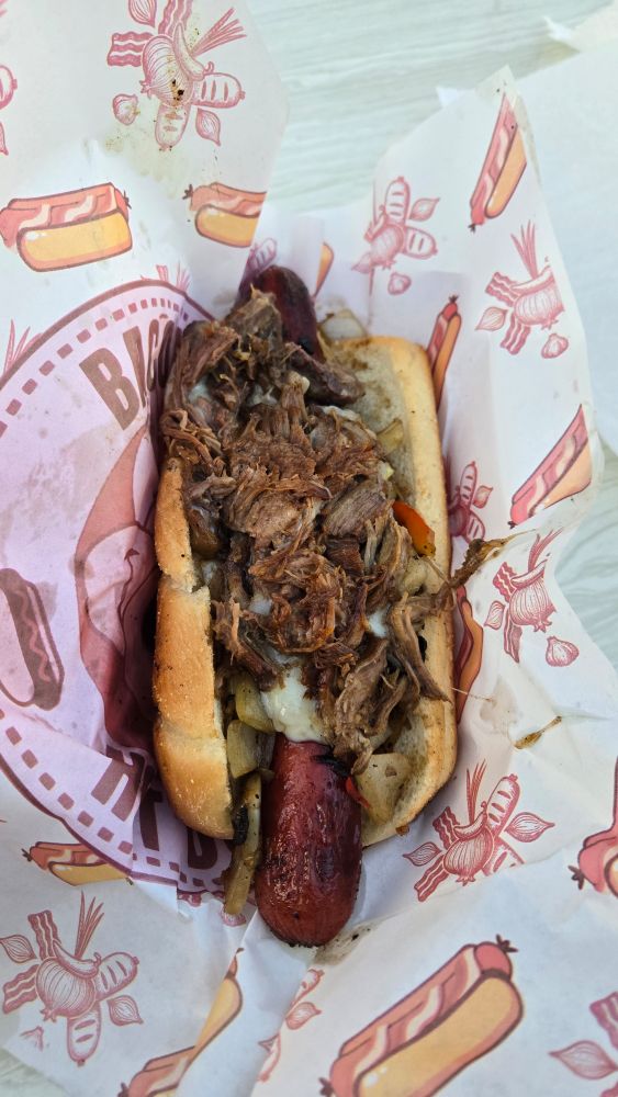 a bacon wrapped hot dog in a bun, topped with grilled onions & peppers, cheese, and shredded beef