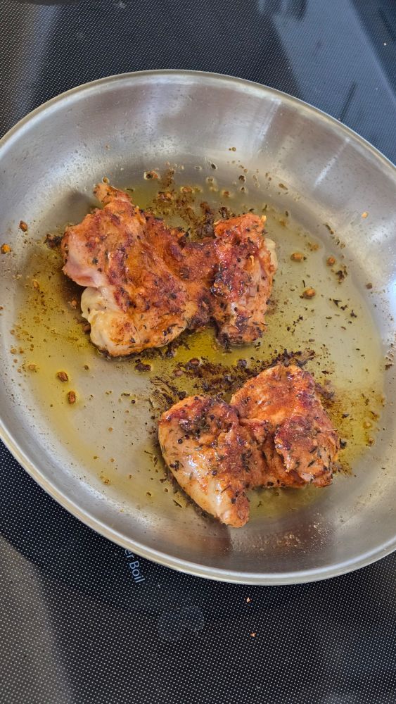 two chicken thighs cookinh on the stove in a steel pan with seed oils. perfectly browned on top with visible hot honey rub.