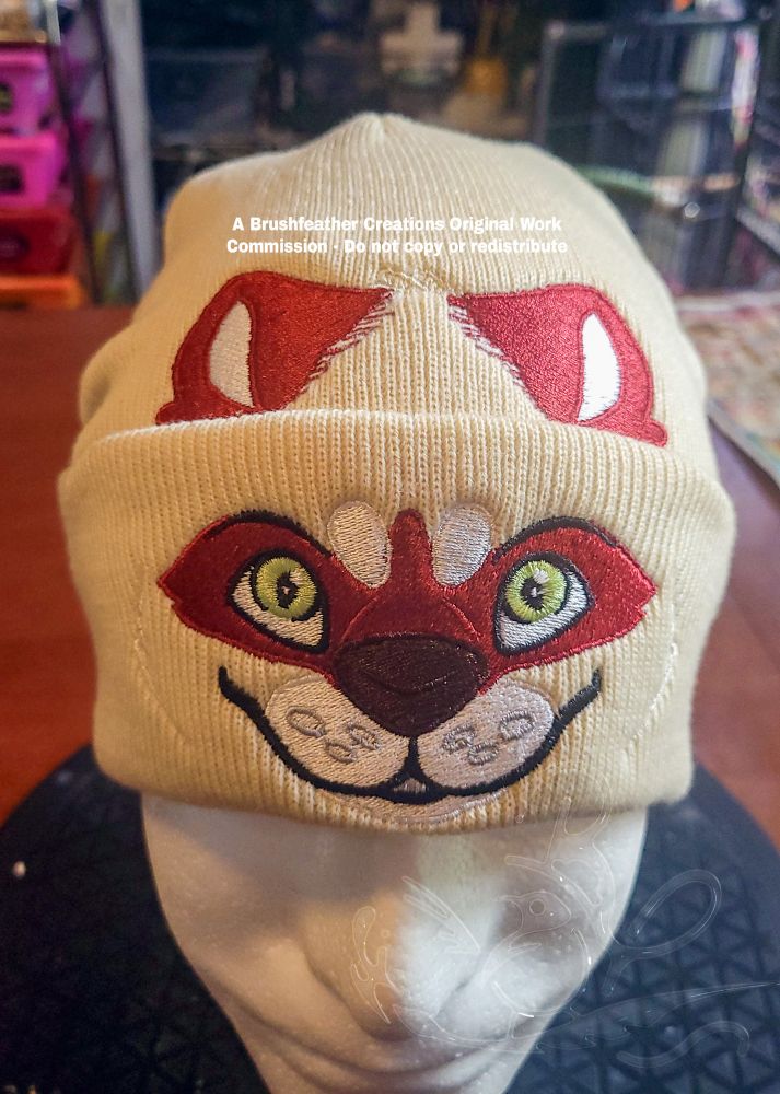 A cream colored hat with a red and tan otter/raccoon character embroidered on the cuff and behind the cuff. 