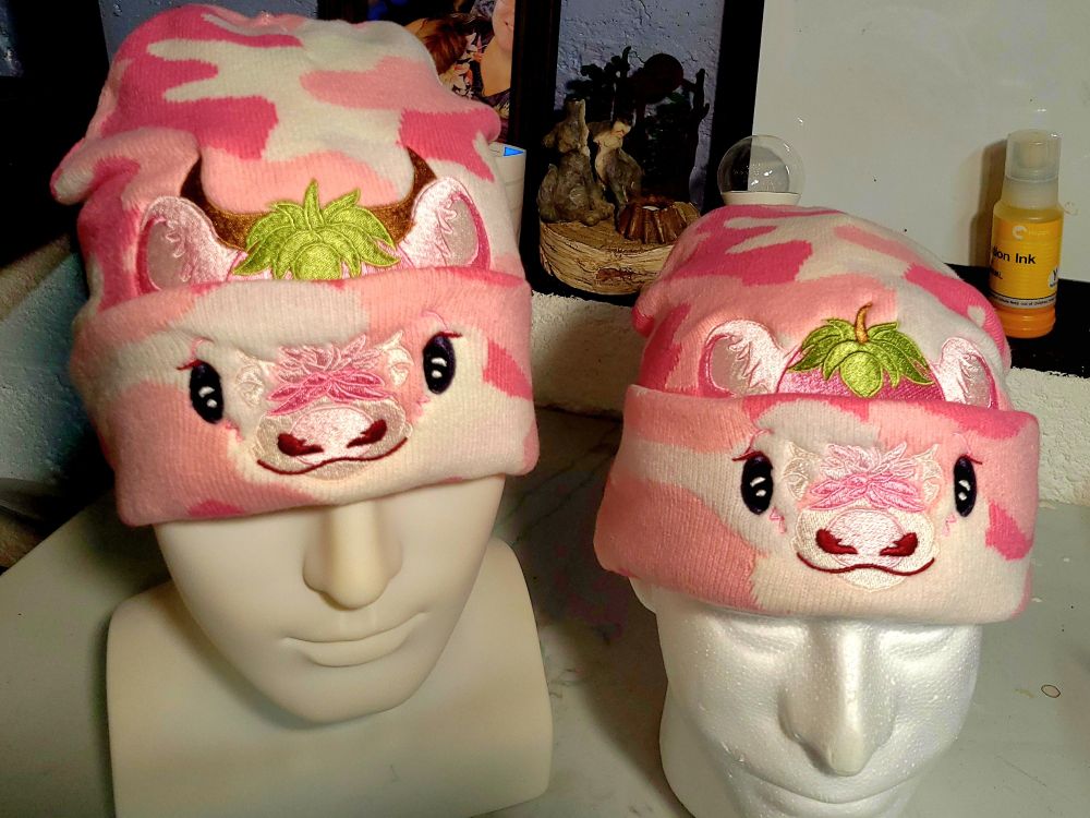 Two pink camo hats with a strawberry cow and strawberry bull design embroidered on and behind the cuff. 