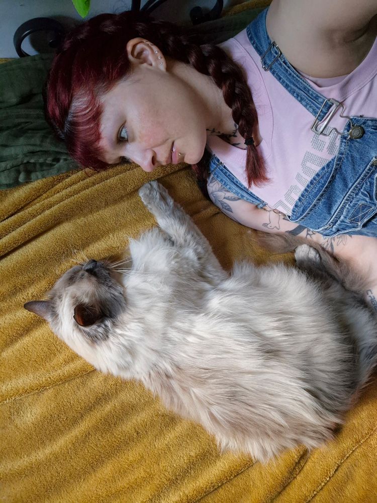me, a white woman with burgandy hair in braids, laying on the bed next to toby, my bluepoint ragdoll cat. one front paw is stretched out to my face