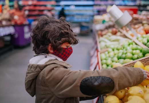 A child in a mask selecting fresh produce at a grocery store.
