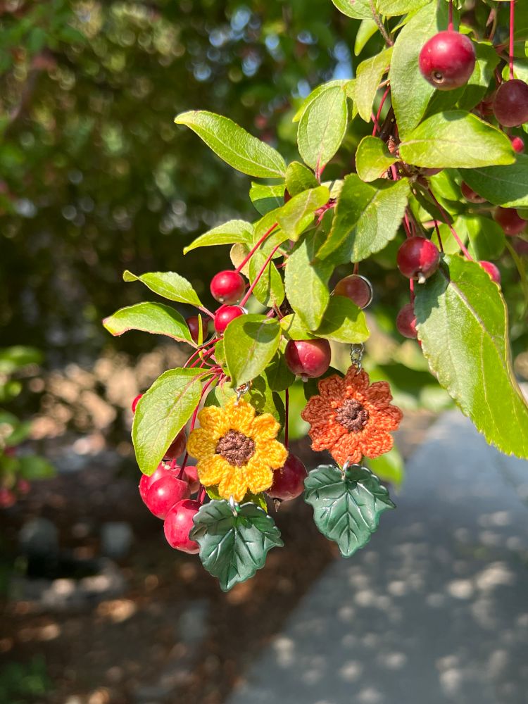 Sunflower crochet earrings one in yellow and one in dark orange. They each have a dark green leaf charm and are hanging in my crabapple tree.