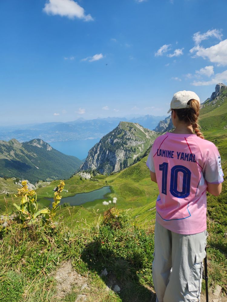 Une jeune femme en maillot du barça contemple un paysage de montagne un petit lac en contre bas et une ouverture sur un grand lac dans la vallée