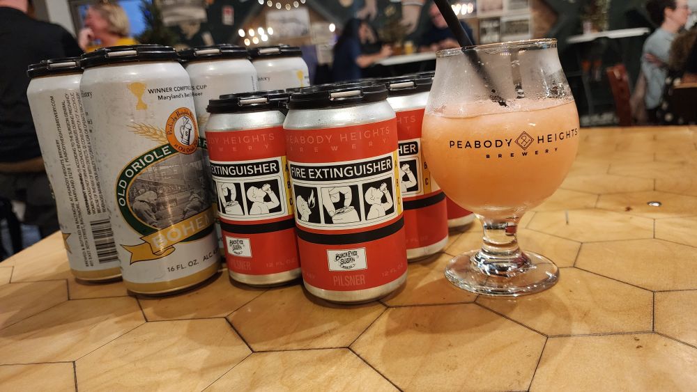 Two six packs and a goblet of mead slush sit stop a hexagonal table inside a brewery tap room