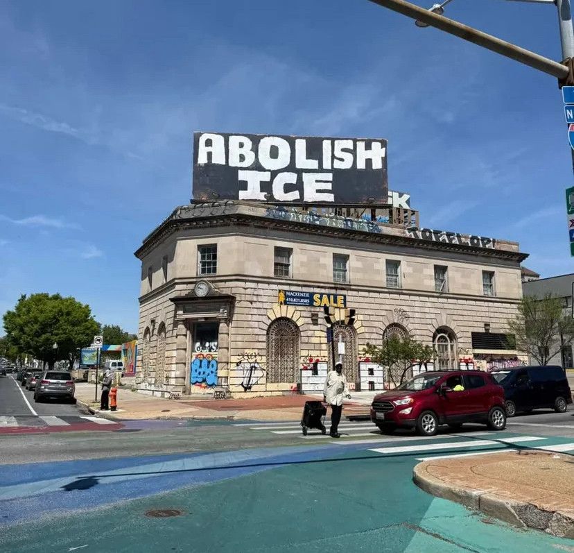 A billboard on top of a building on the corner of Baltimore's North Ave and Howard Street, reading "ABOLISH ICE" in hand-painted white letters on a black background 