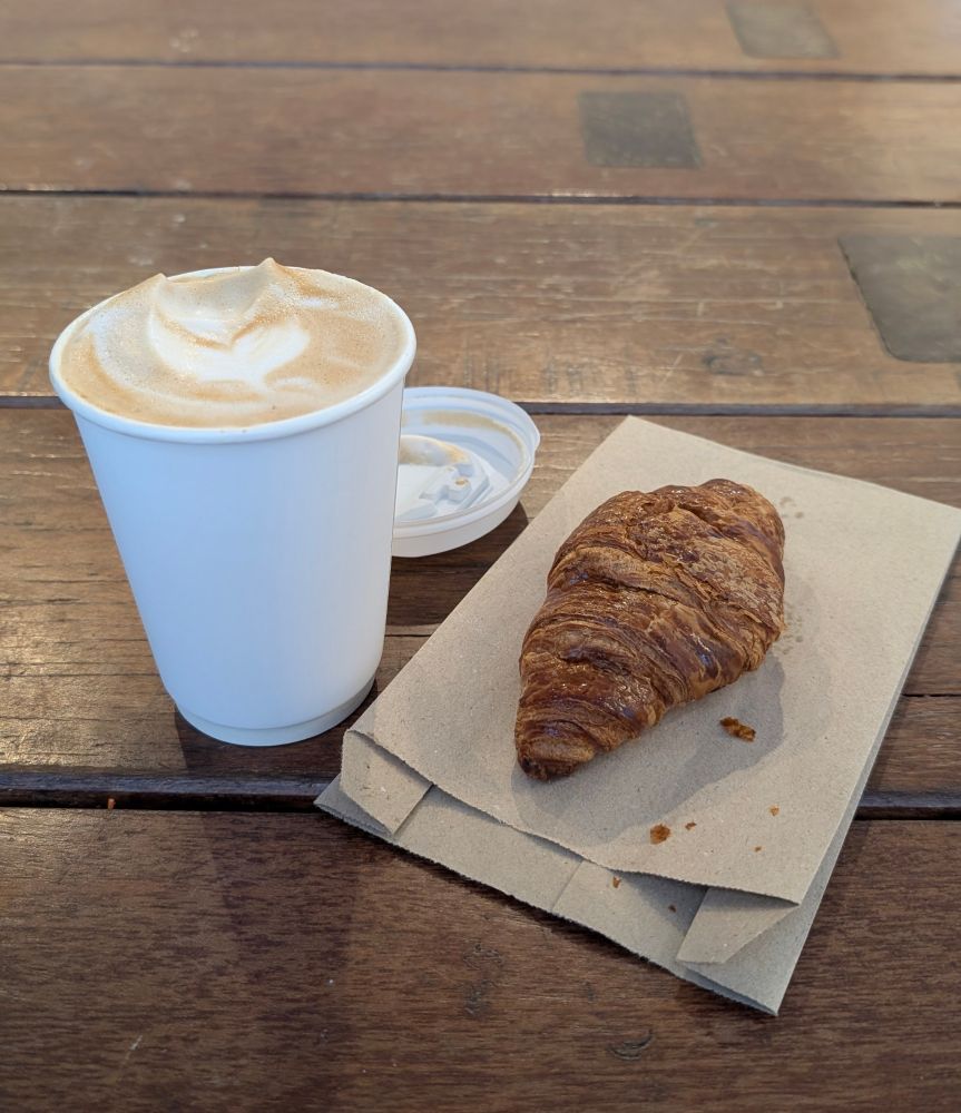 A cup of coffee and a croissant. The foam on the coffee looks like extended devil horns 