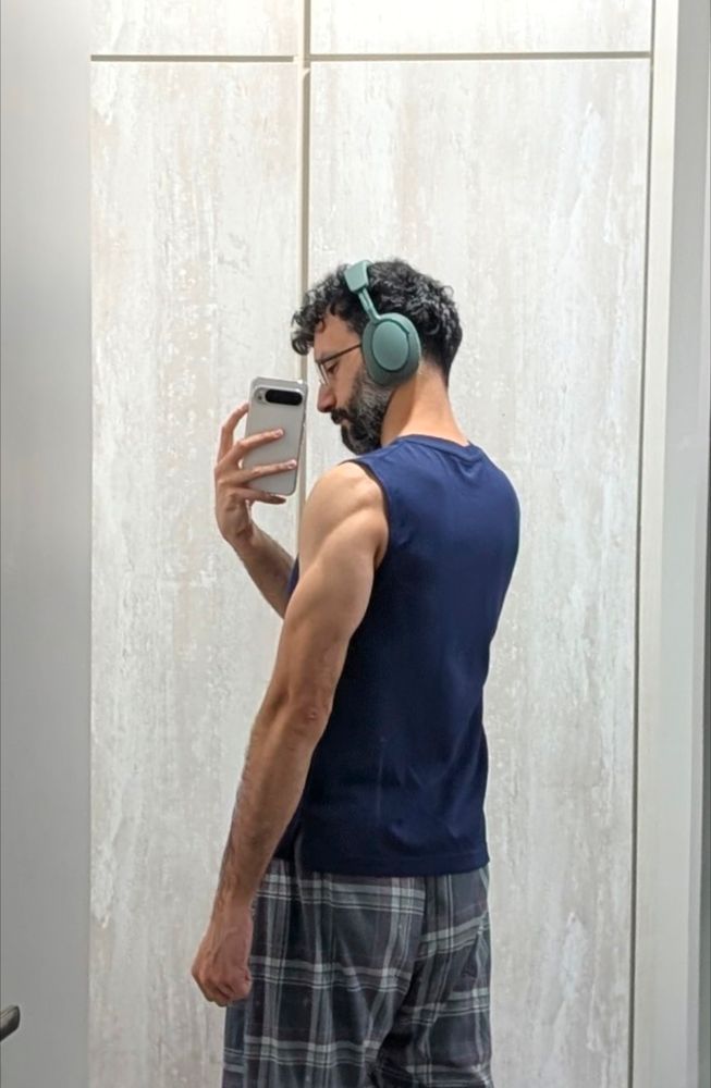 A mid-30s bearded bespectacled man taking a picture of his back. He's wearing a blue tank top with arms exposed. He's wearing green headphones 