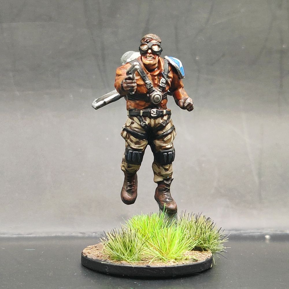 A pulp hero in a red leather jacket, pale trousers and black and gold goggles. He's holding a silver pistol and has a blue shoulder pad with a white star on it.