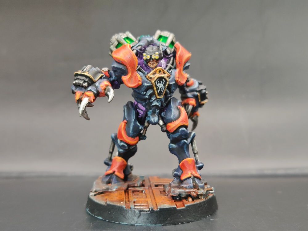 A heavily armoured hunter in dark blue armour with orange panels. He wears a purple undersuits, has glowing yellow eye lenses and two glowing green capacitors on his back.