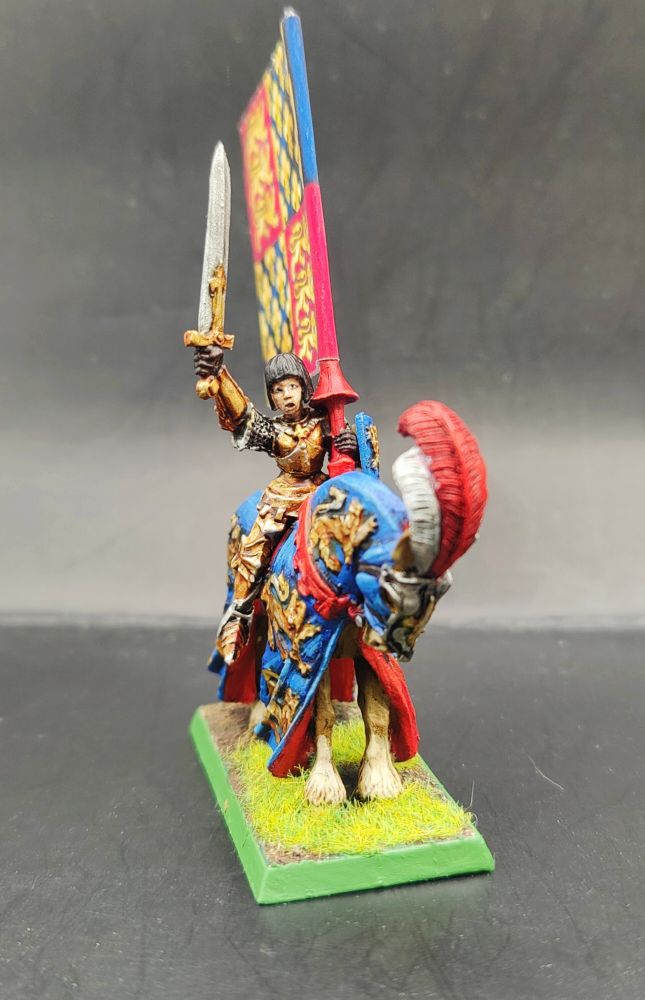 A young girl in golden armour holding a sword aloft whilst riding on a horse wearing a blue and red caprison with golden lions on it. She is also holding a banner.