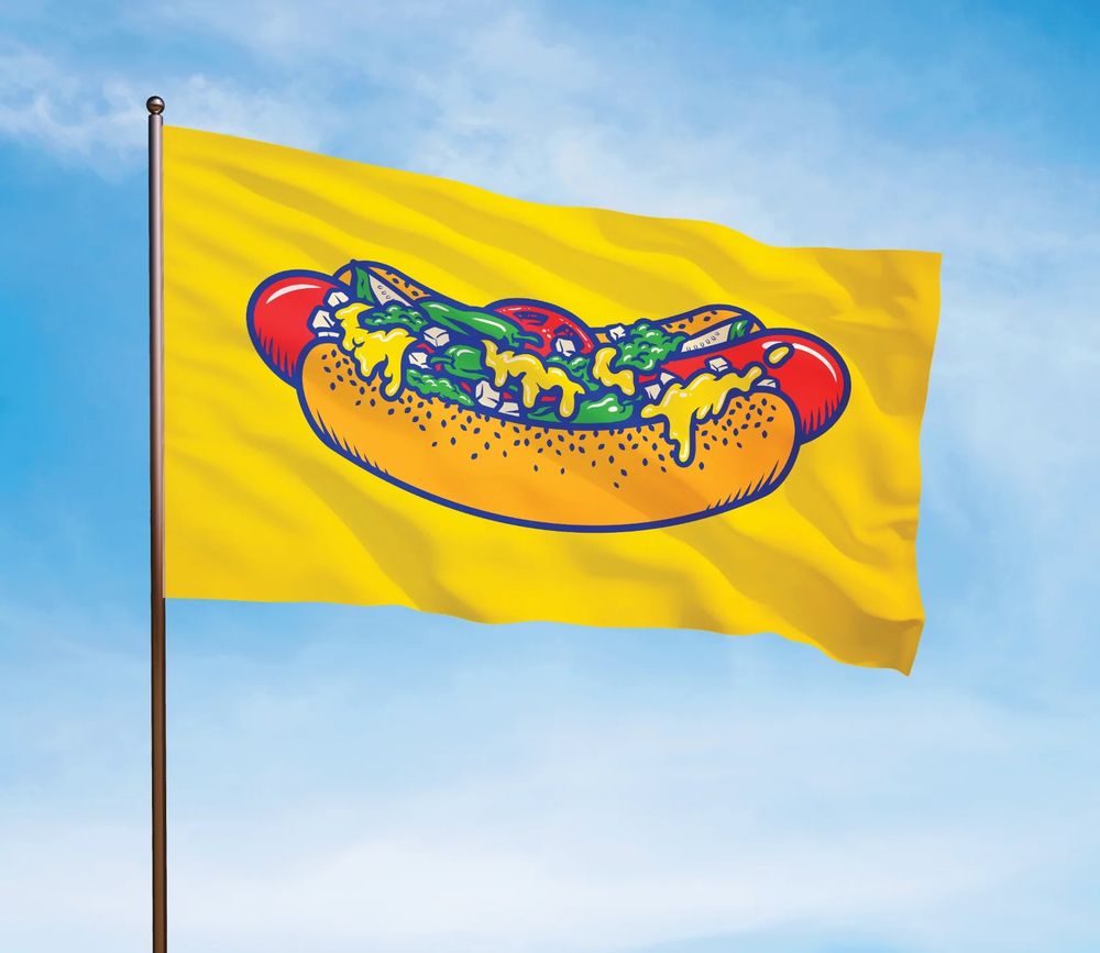 A yellow flag with a Chicago style hot dog graphic flies in front of a blue sky