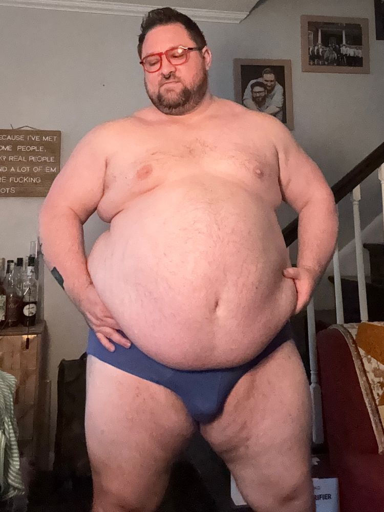 Man in dark blue briefs and red glasses, holding his belly and looking down at it with a grin on his face. A letter board in the background reads “because I’ve met some people, OK? Real people. And a lot of ‘em are fucking idiots.”