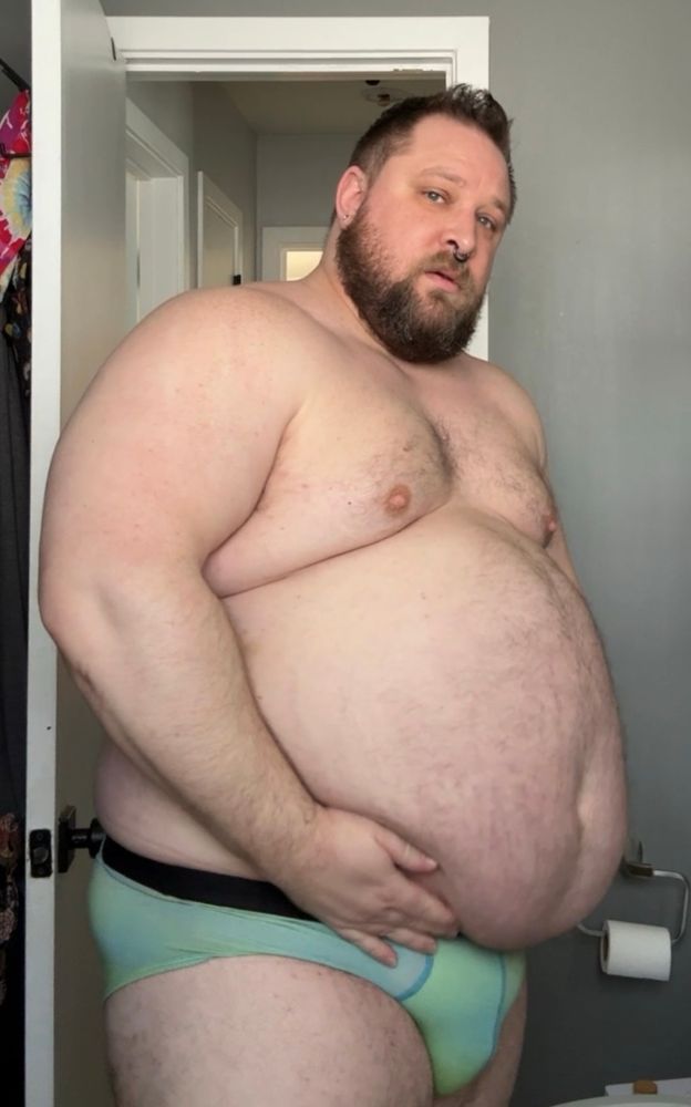 A rotund man in blue-green briefs holding his belly 
