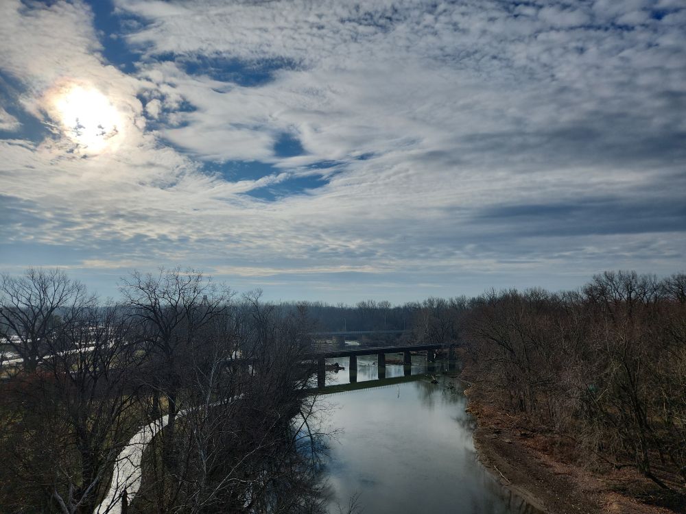 photo of a river cutting through the trees from up high. The trees have no leaves. A walking path snakes between them on the left, following the river. They sky above is very cloudy, but blue peaks through in a few places.