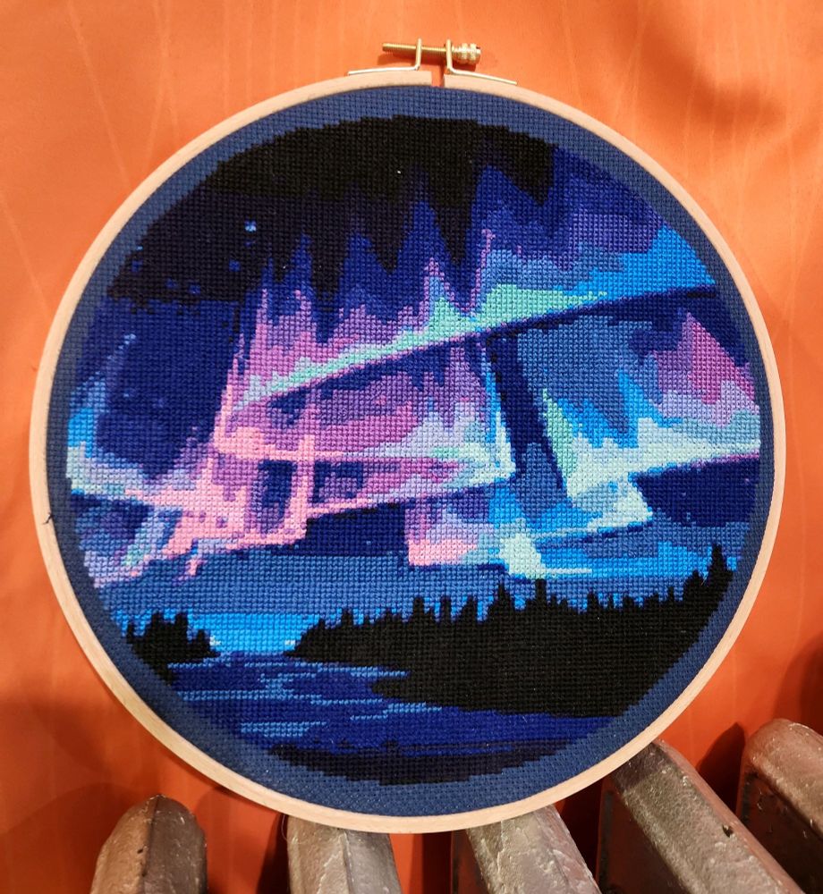 A cross stitch project of the aurora borealis in a plain wood hoop. It is a field of siwlring blues, purples, and pinks over a dark forest and lake.