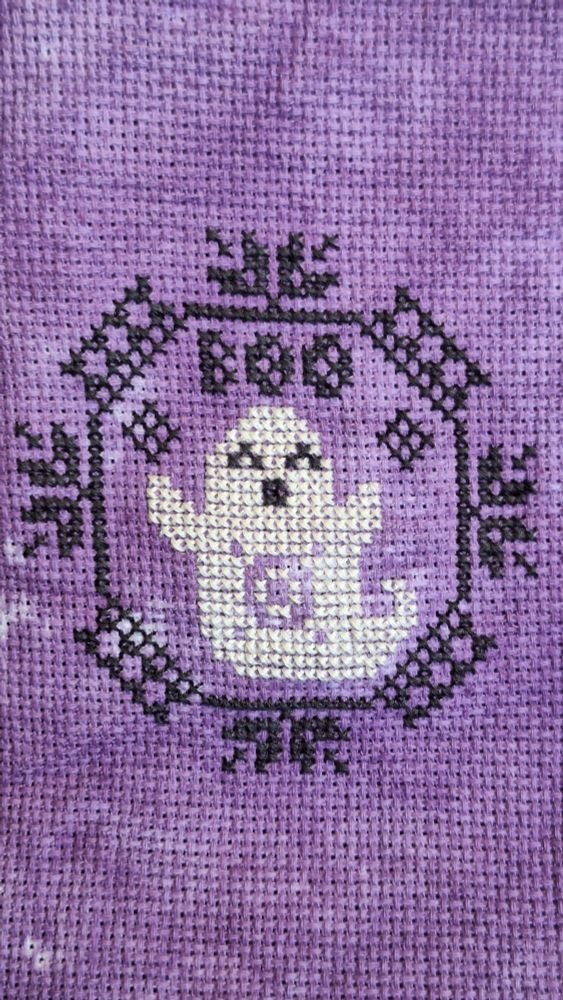 A cross stitch piece of a quaker motif with a ghost in the middle and BOO on top of Ghost 